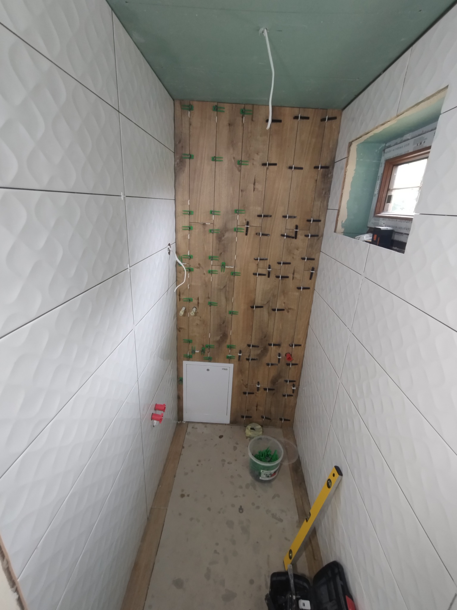 Ściany łazienki w trakcie układania białych płytek z falistym wzorem, fragment drewnianej ściany z krzyżykami dystansowymi, poziomica i wiadro z zielonymi klinami na podłodze.