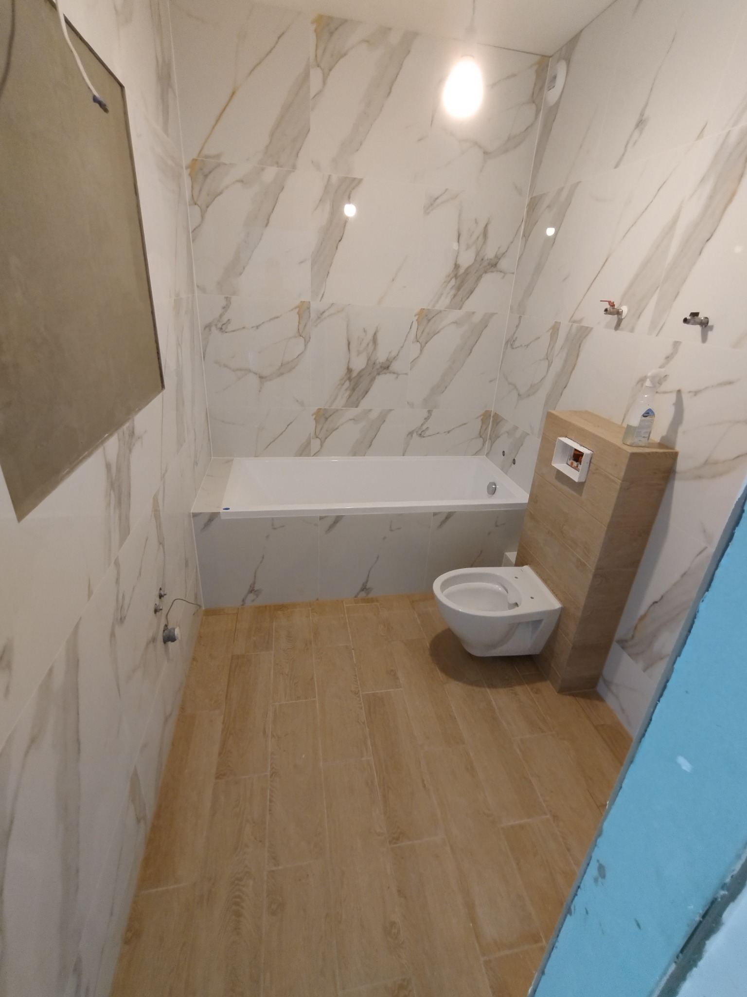 Nowoczesna łazienka z marmurowymi płytkami na ścianach, drewnopodobną podłogą, białą wanną i toaletą ze spłuczką w zabudowie z drewnianych paneli. Na zabudowie spłuczki stoi butelka płynu.