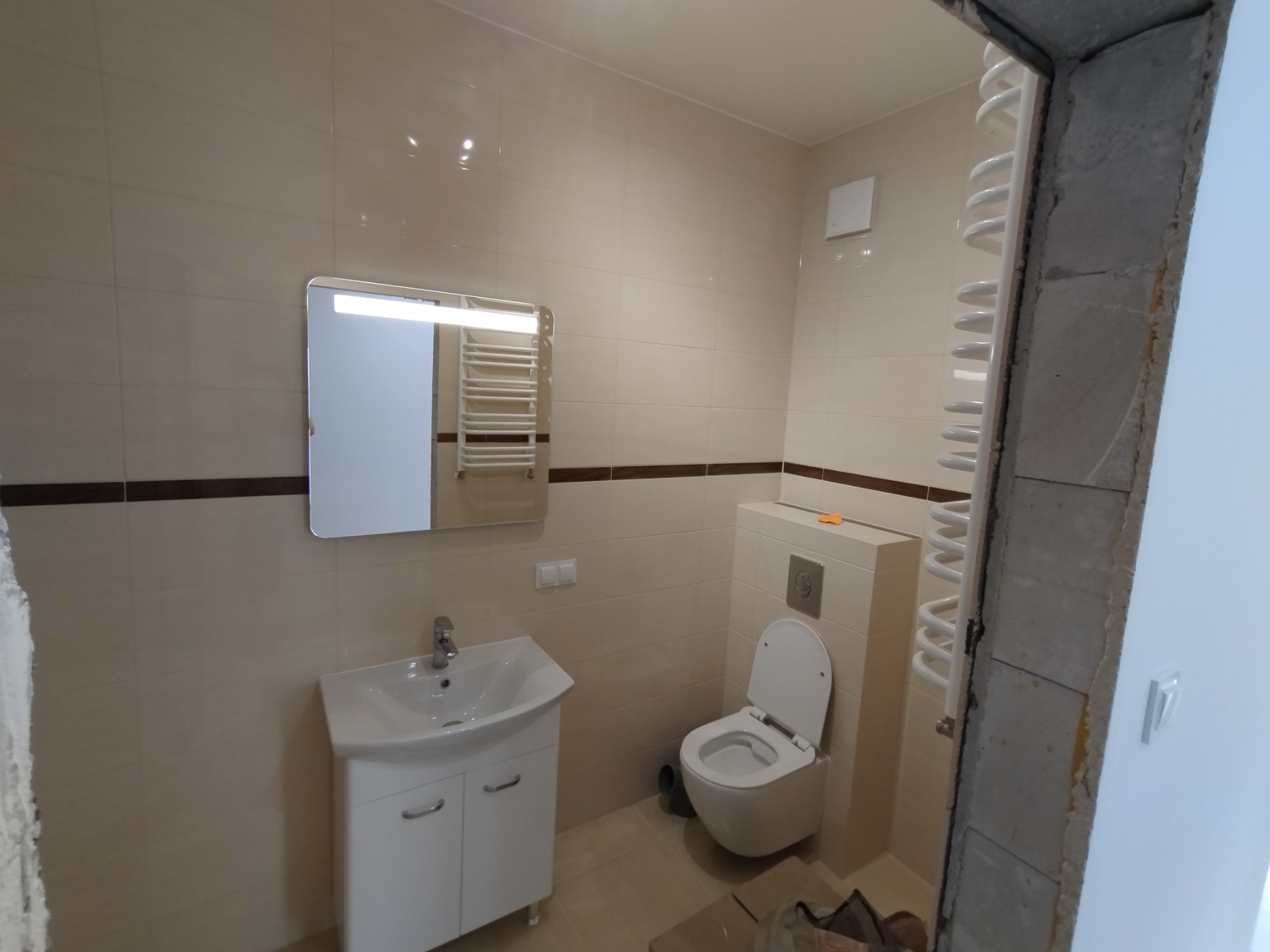 Wykończona łazienka z beżowymi płytkami, białą umywalką z szafką, toaletą i grzejnikiem drabinkowym, widoczne surowe wykończenie framugi drzwi.