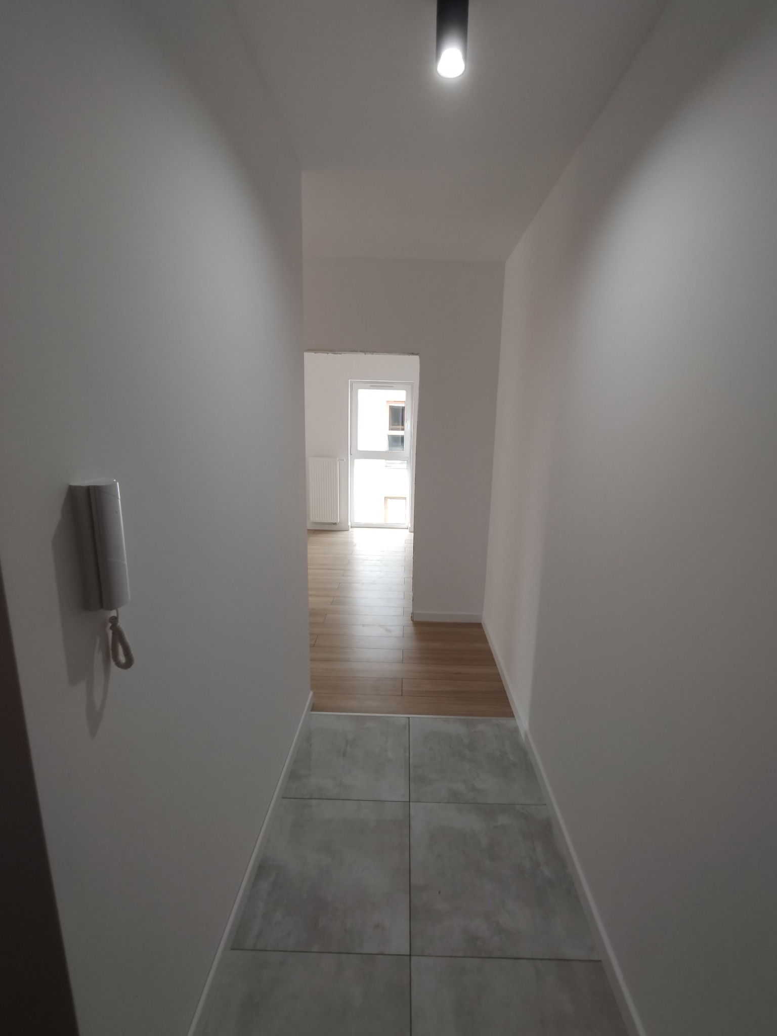 Wnętrze korytarza po remoncie: szare płytki podłogowe przechodzące w drewnopodobne panele, białe ściany z domofonem, oświetlenie sufitowe punktowe.