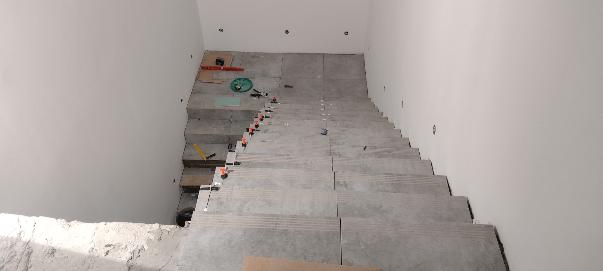 Kładzenie szarych płytek na schodach wewnętrznych z użyciem poziomowania, widoczne narzędzia glazurnicze i niedokończone ściany.