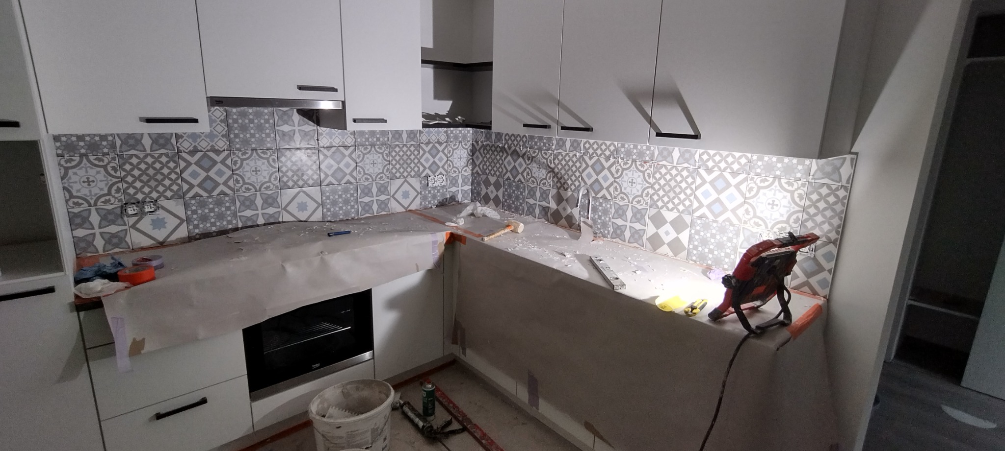 Montaż wzorzystych, szaro-białych płytek na ścianie w kuchni, widoczne narzędzia pracy, poziomica, lampa halogenowa, zabezpieczony blat, białe szafki kuchenne z czarnymi uchwytami.