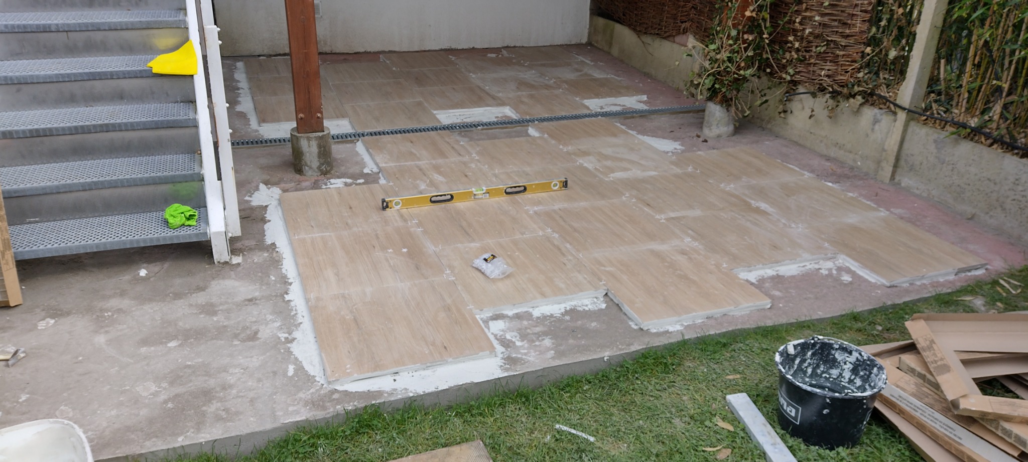 Układanie płytek imitujących drewno na zewnątrz, widoczna poziomica, wiadro z zaprawą, schody metalowe i trawa.