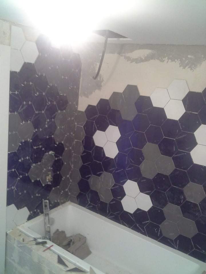 Układanie heksagonalnych płytek w odcieniach bieli, szarości i granatu na ścianie w łazience nad wanną, widoczny fragment surowego tynku i wystający kabel.