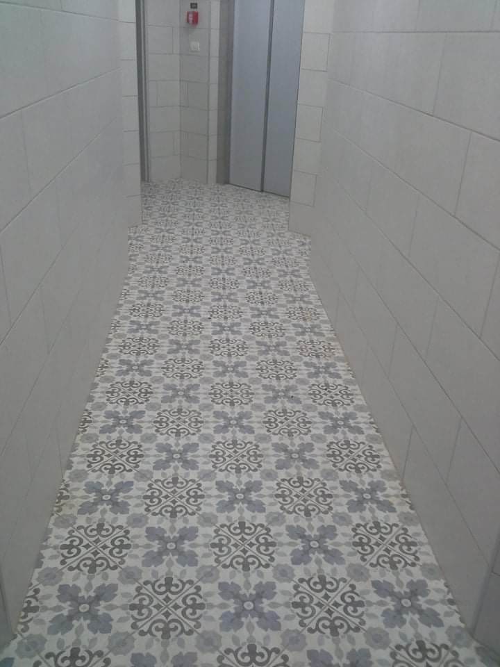 Wąski korytarz wyłożony jasnymi, wzorzystymi płytkami podłogowymi w odcieniach szarości i bieli, ściany wyłożone prostokątnymi, białymi płytkami, w tle widoczne drzwi.