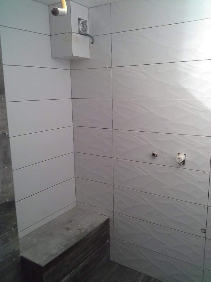 Ściany łazienki w trakcie układania białych, poziomych płytek z delikatnym wzorem falistym; widoczne krzyżyki dystansowe, fragment obudowy rur i przygotowane podejścia wodne.