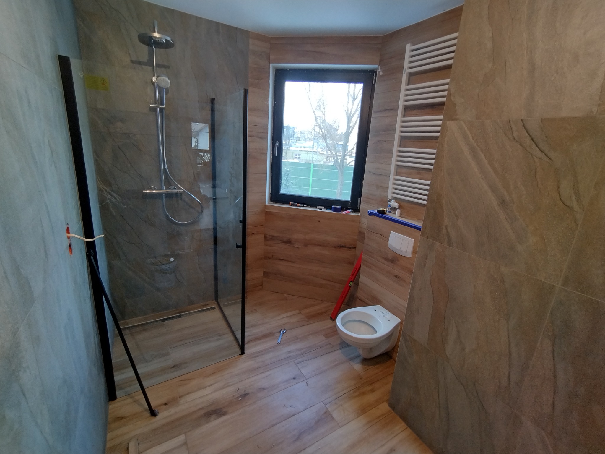 Nowoczesna łazienka w trakcie wykańczania: kabina prysznicowa typu walk-in z czarnymi profilami, wisząca toaleta, grzejnik drabinkowy, płytki imitujące drewno na ścianach i podłodze, narzędzia...