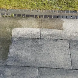 Częściowo oczyszczona, szara kostka brukowa przy granicy trawnika, widoczne różnice w kolorze i zabrudzeniu przed i po czyszczeniu.