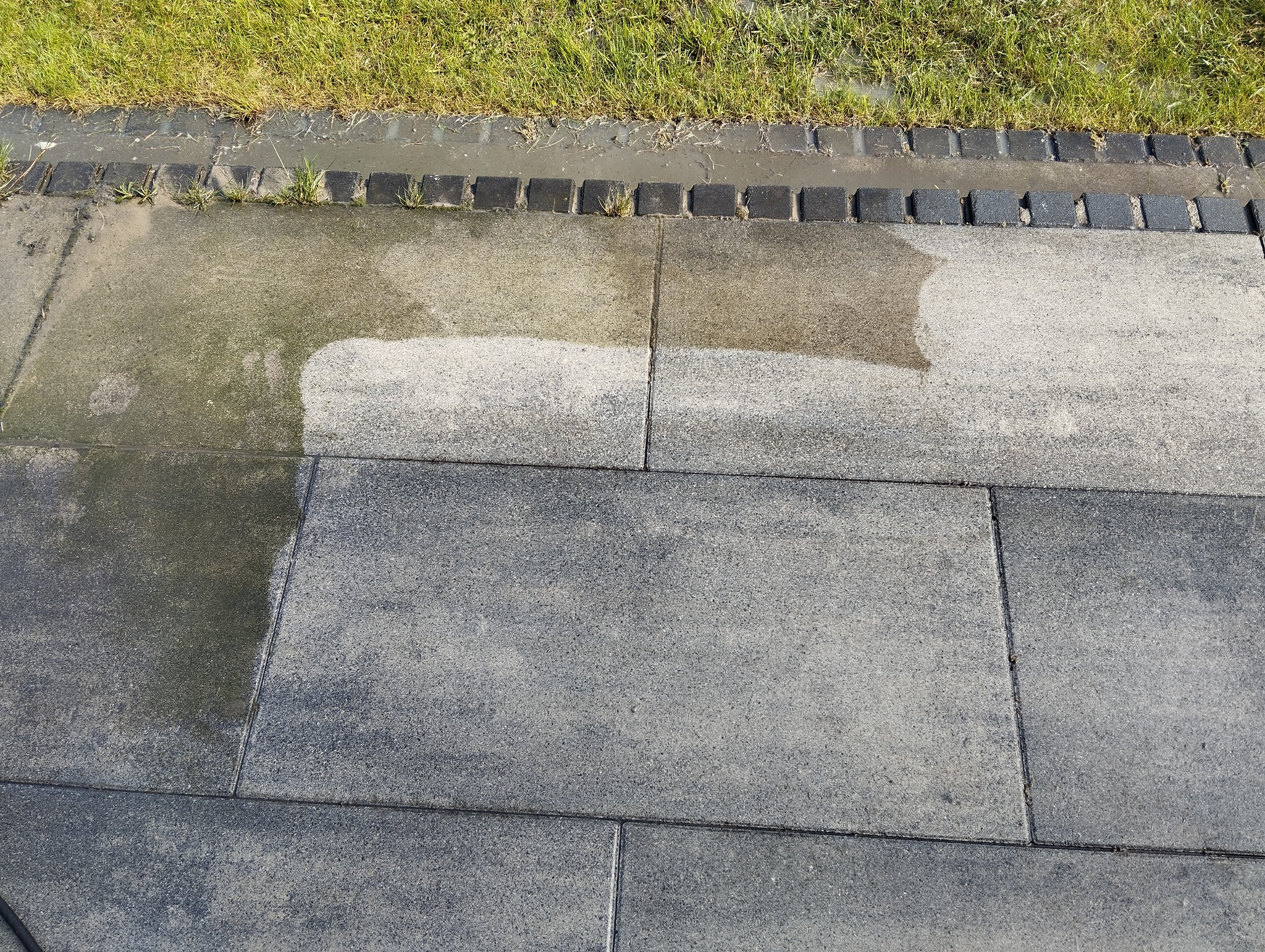 Częściowo oczyszczona, szara kostka brukowa przy granicy trawnika, widoczne różnice w kolorze i zabrudzeniu przed i po czyszczeniu.