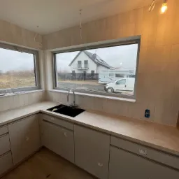 Wnętrze łazienki w trakcie remontu: widok na umywalkę z baterią, blat, okno z widokiem na dom i zaparkowany samochód dostawczy, oraz fragment ściany z ułożonymi płytkami i wystającym kablem...