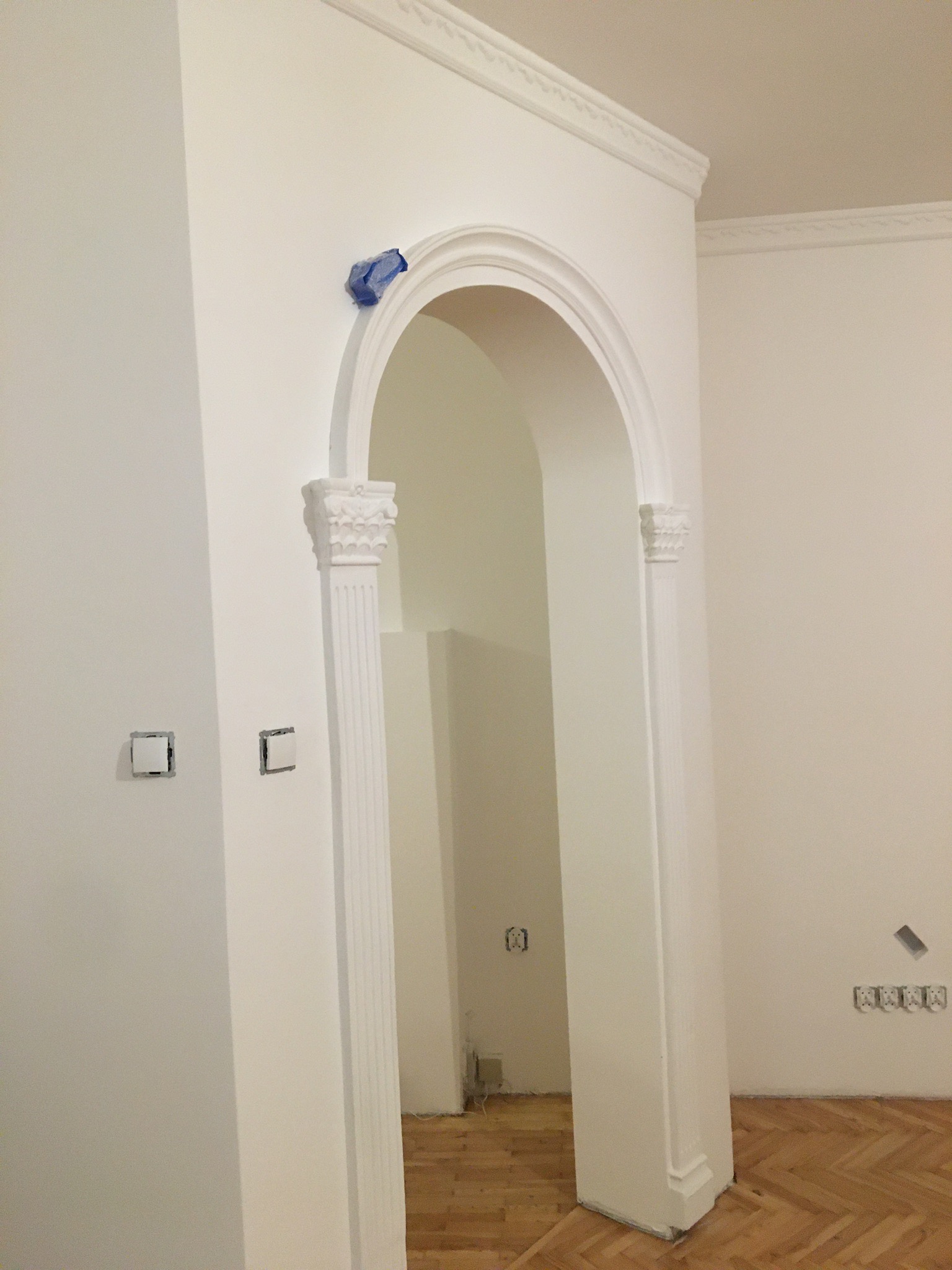 Białe, odnowione wnętrze z ozdobnym łukiem, listwami przysufitowymi i parkietem. Niebieska taśma malarska zabezpiecza górną część łuku. Puszki elektryczne bez osprzętu widoczne na ścianach.