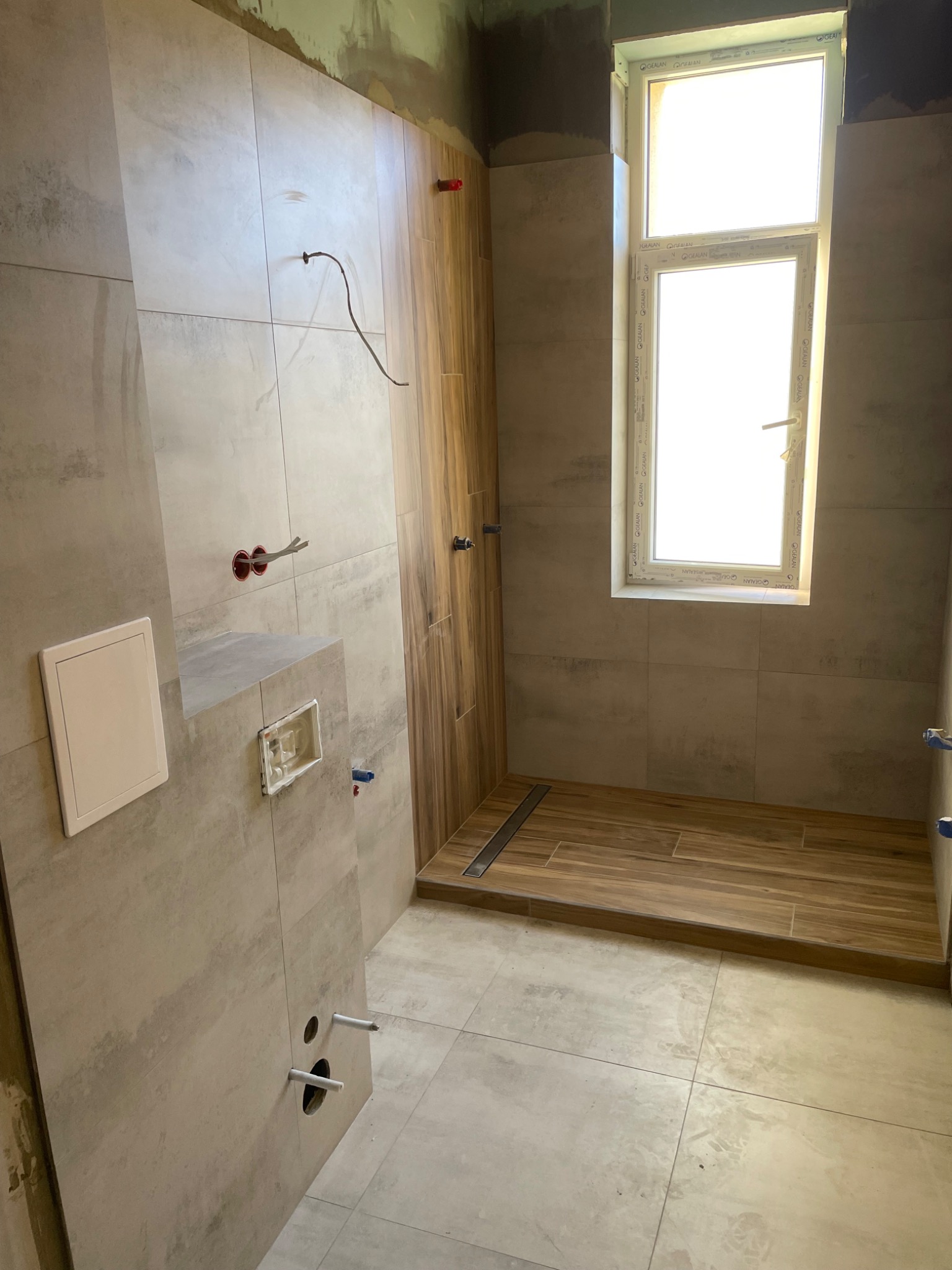 Niedokończona łazienka w trakcie prac: szare płytki imitujące beton na ścianach, drewnopodobne płytki na podłodze w strefie prysznica z odpływem liniowym, widoczne instalacje elektryczne...