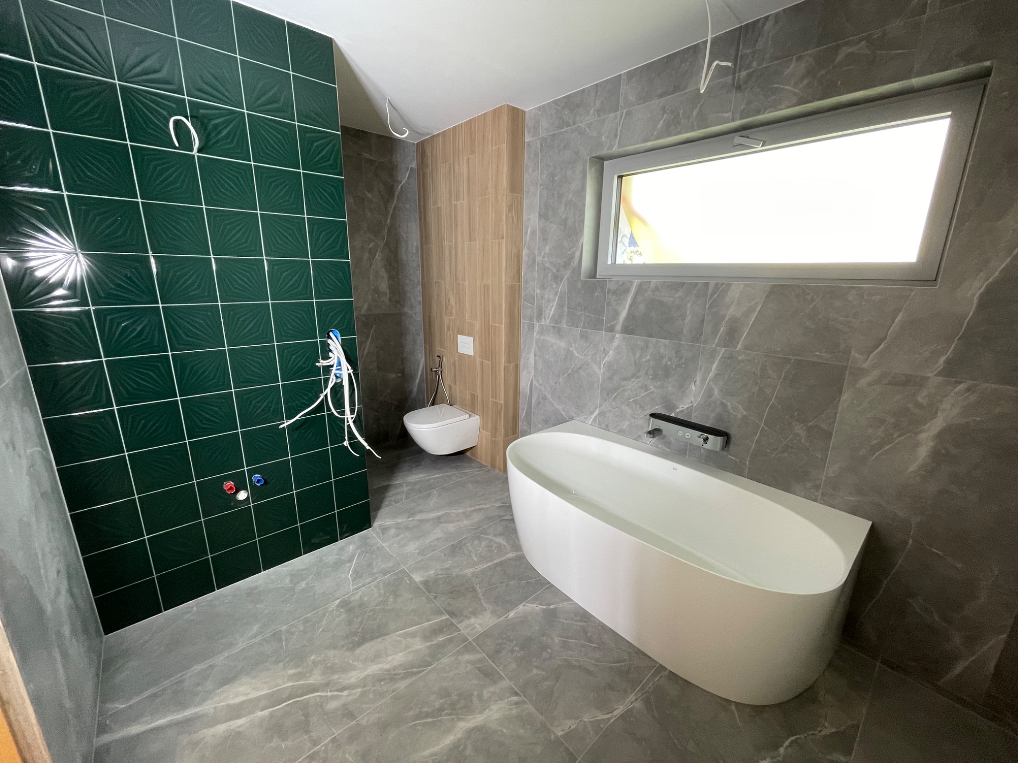 Nowoczesna łazienka w trakcie wykańczania: zielona ściana z wypukłymi kwadratowymi płytkami, marmurowe szare płytki na ścianach i podłodze, wolnostojąca wanna z napełniona wodą, wisząca toaleta...