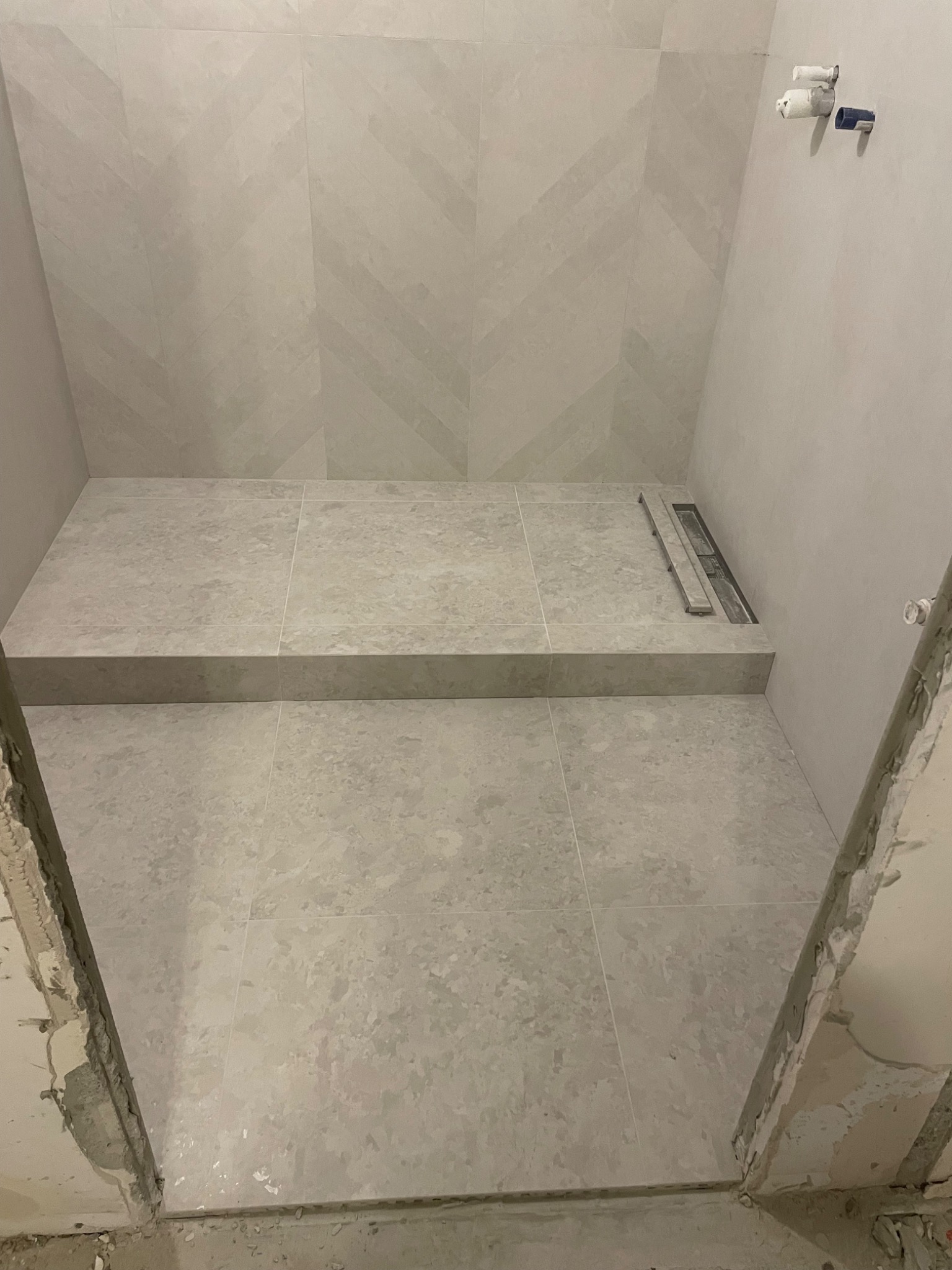 Wykończony brodzik z odpływem liniowym, wyłożony jasnymi, matowymi płytkami w jodełkę na ścianach, widoczne niedokończone obrzeża wejścia do kabiny.