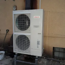 Biała jednostka zewnętrzna klimatyzacji marki Atlantic zamontowana na metalowym stelażu przy ścianie budynku, obok widoczna łopata i wiadro.