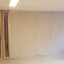 Wnętrze w trakcie prac wykończeniowych: ściana z płyt kartonowo-gipsowych z widocznymi łączeniami, drewniana konstrukcja ościeżnicy w tle, narzędzia budowlane przy ścianie, w tym ukośnica.