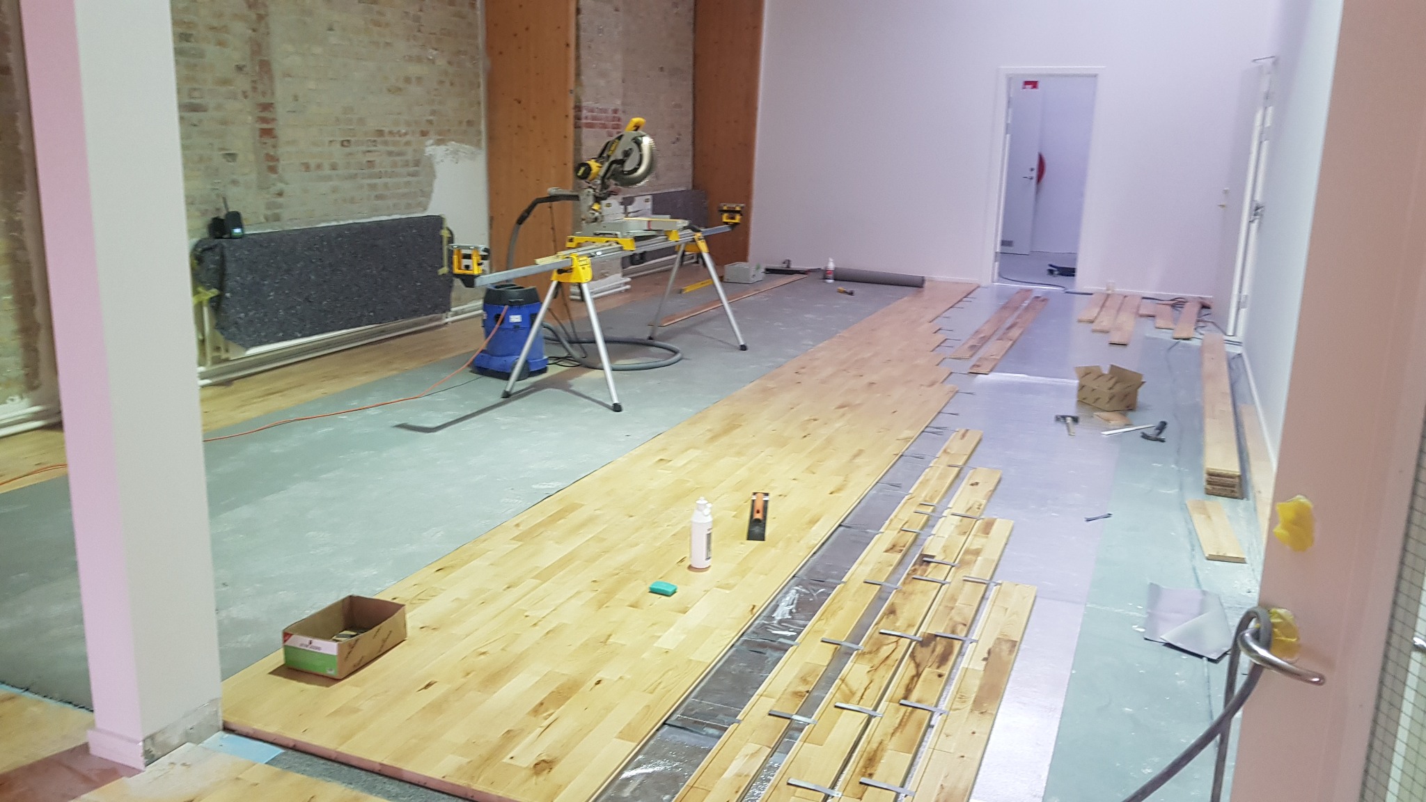 Montaż drewnianej podłogi w trakcie prac: widoczna ukończona część parkietu, nieułożone deski, piła elektryczna na stojaku, odkurzacz przemysłowy, narzędzia oraz klej na podłodze, surowa ściana...