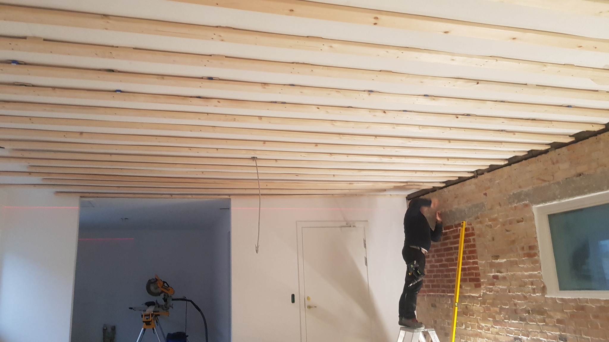 Montaż drewnianej konstrukcji sufitu z widoczną czerwoną linią lasera, pracownik na drabinie mierzy ścianę z cegły żółtą poziomicą, w tle białe ściany i drzwi.