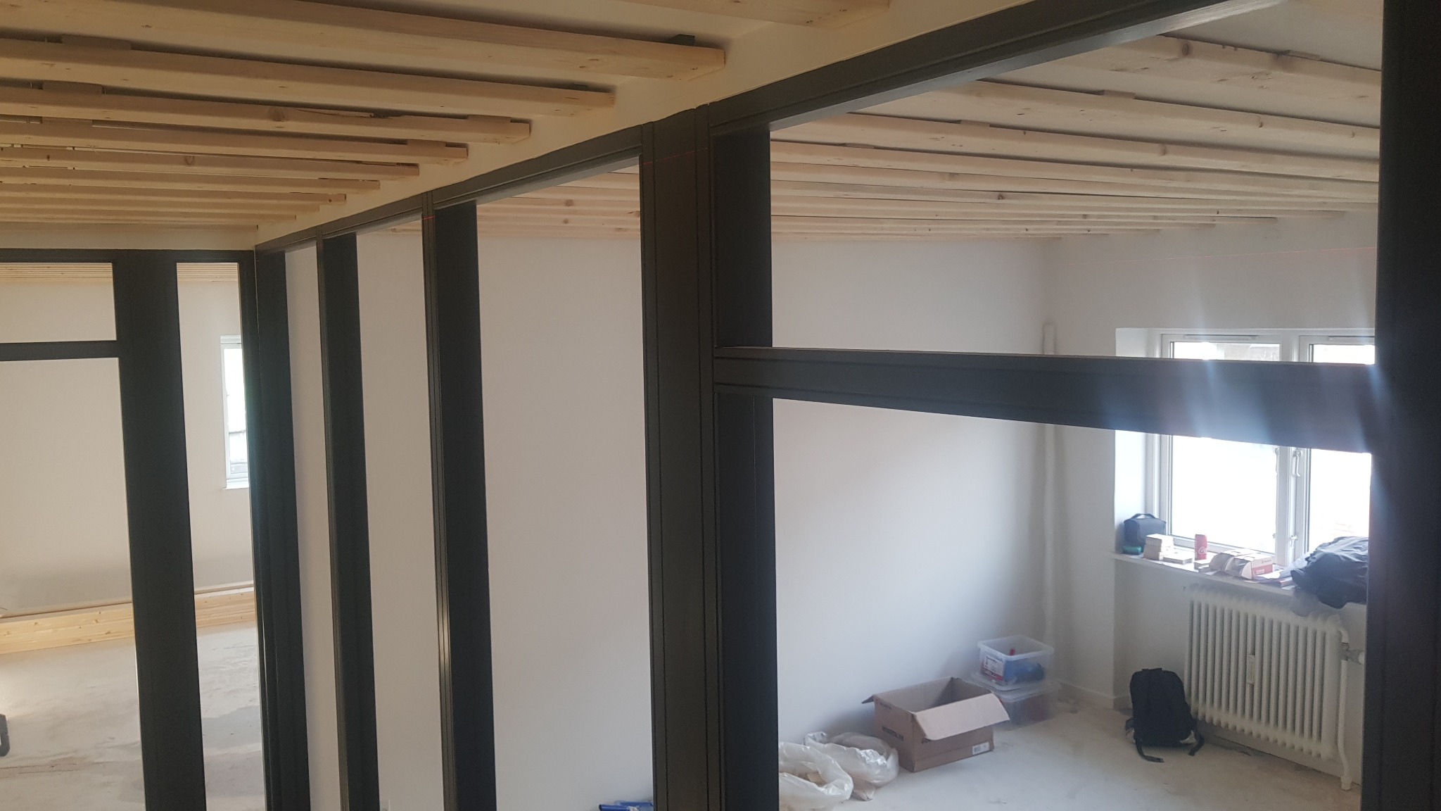 Konstrukcja ścian działowych z czarnymi profilami w trakcie montażu, widoczny drewniany szkielet sufitu i surowe, białe ściany w tle.