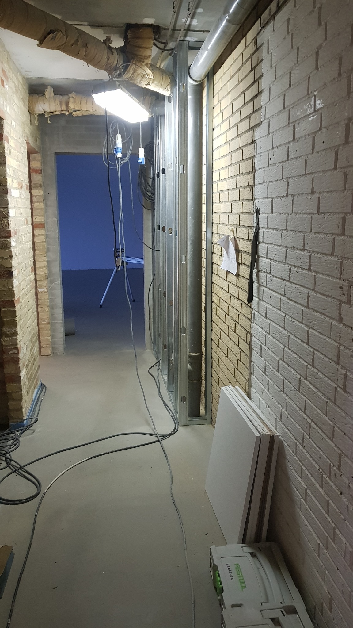 Korytarz w trakcie prac wykończeniowych: widoczne profile metalowe do montażu ścianek działowych, częściowo pomalowana ceglana ściana, płyty gipsowo-kartonowe oparte o ścianę i skrzynka narzędziowa...