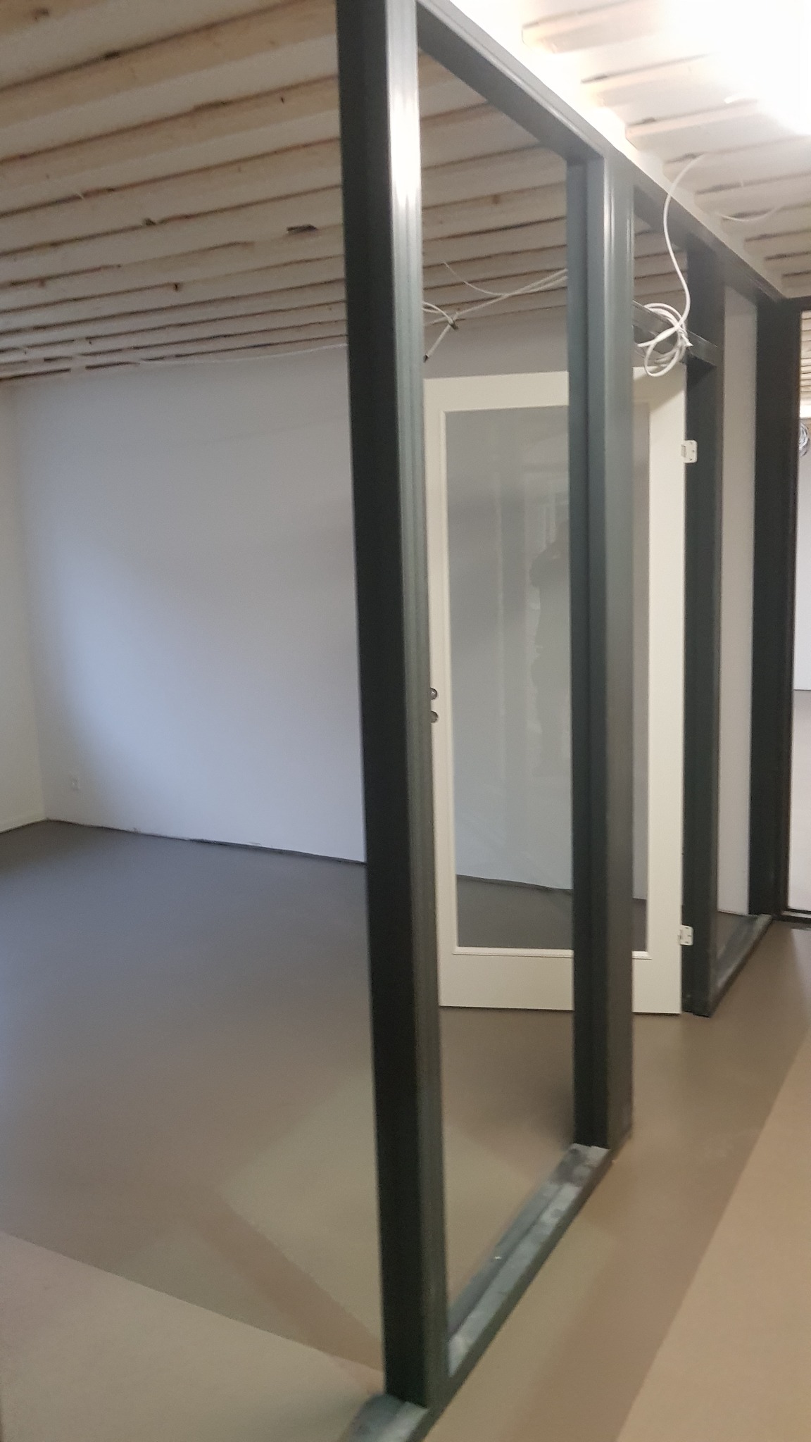 Nowo zamontowane, białe drzwi z przeszklonymi panelami, osadzone w ciemnoszarej, metalowej ramie w surowym wnętrzu z drewnianym stropem i gładką, szarą podłogą.