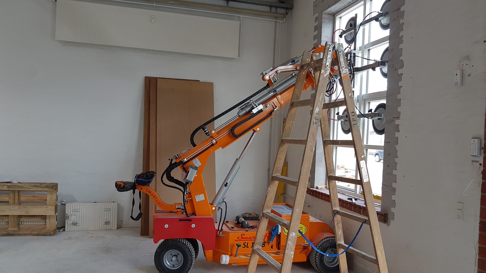 Pomarańczowy podnośnik próżniowy Smartlift SL180 z przyssawkami zamontowany na oknie podczas montażu szyby, obok drabina drewniana, w tle białe ściany i palety.