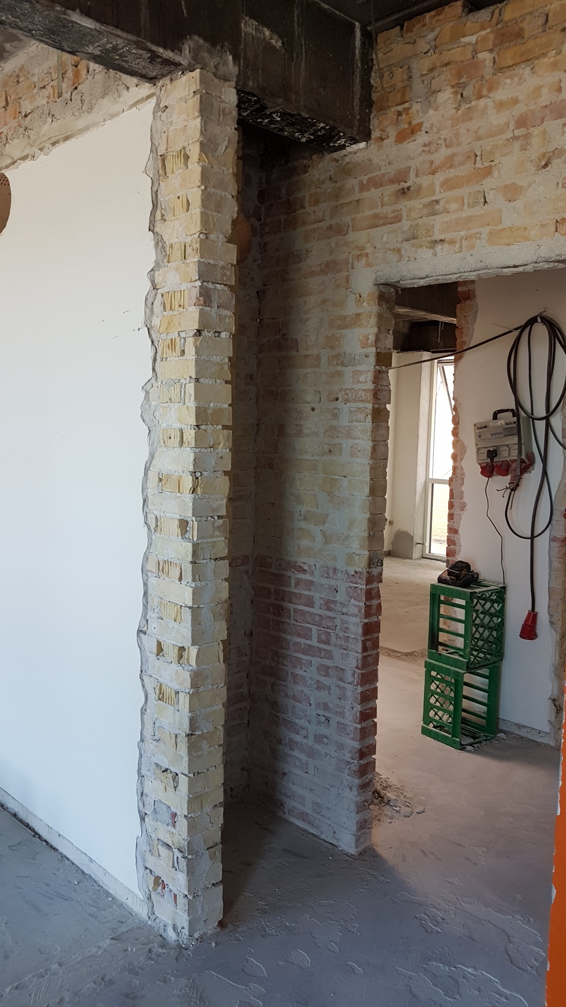 Wnętrze w trakcie prac murarskich i tynkarskich; surowa cegła, fragment ściany pokryty tynkiem, widoczne ślady po kuciu, w tle skrzynki i przedłużacz.