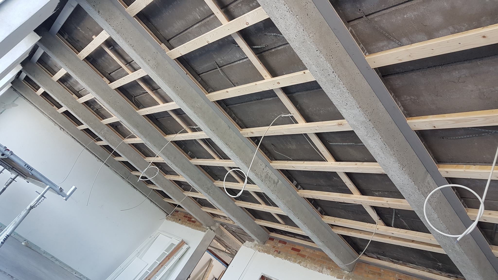 Betonowe belki stropowe z drewnianą konstrukcją podwieszaną, widoczne przewody elektryczne, surowy stan wykończenia wnętrza.