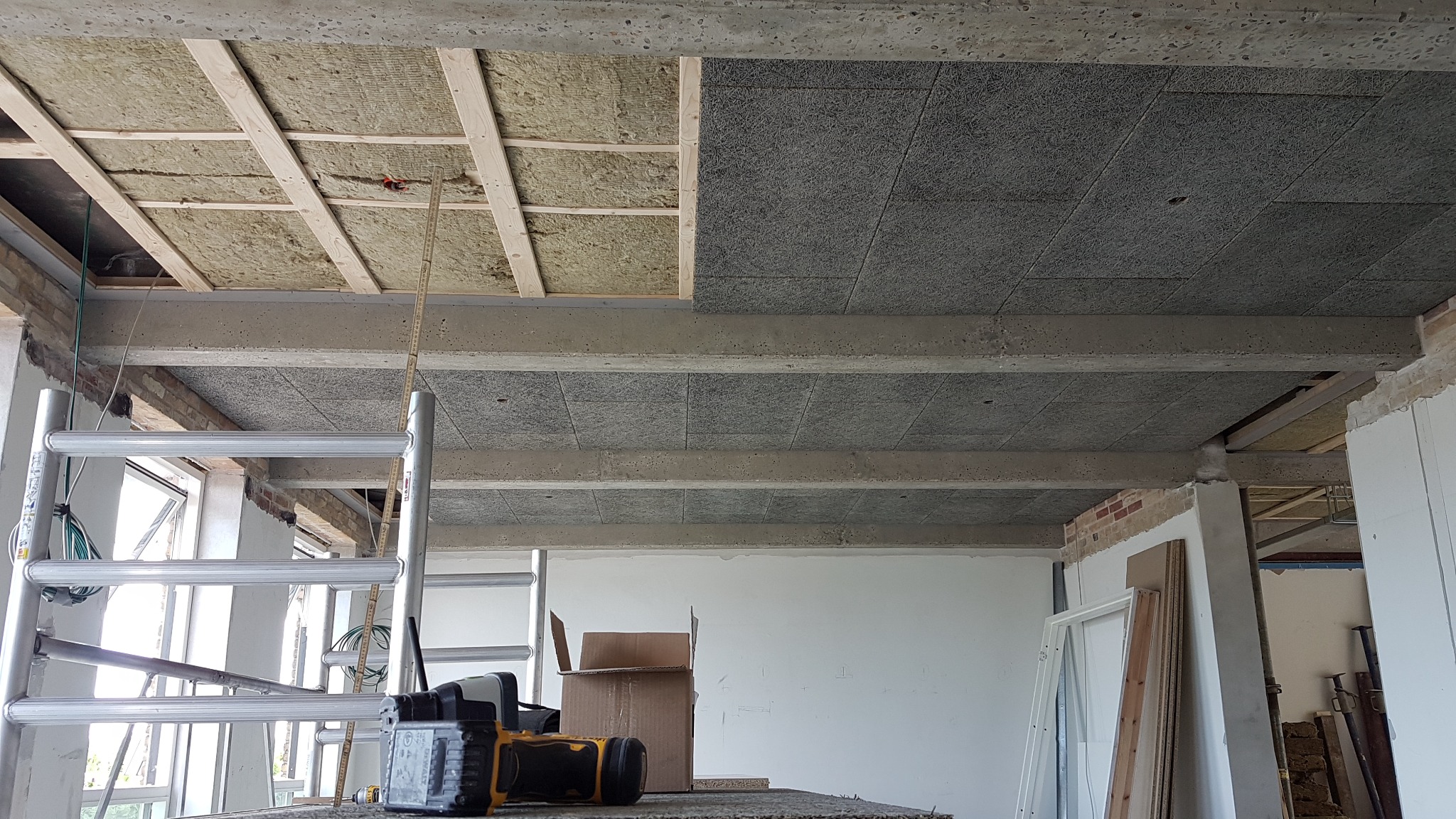 Montaż sufitu podwieszanego z izolacją, widoczna konstrukcja drewniana, wełna mineralna i płyty akustyczne, drabina aluminiowa i narzędzia na pierwszym planie.