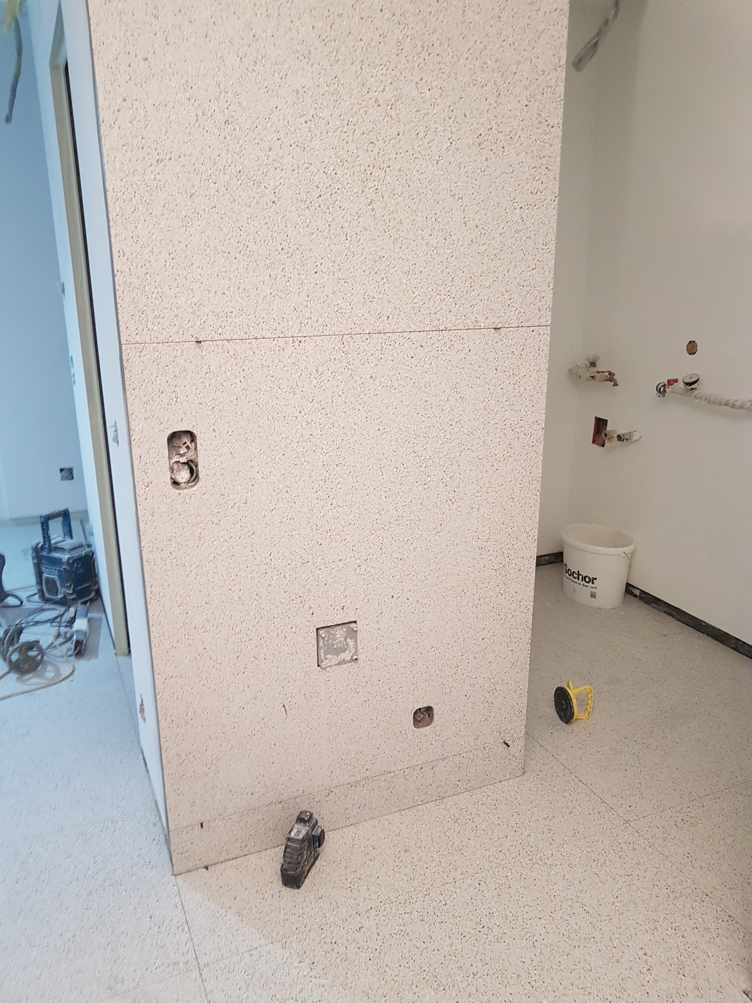 Montaż płyty o porowatej strukturze z widocznymi puszkami elektrycznymi i narzędziami na podłodze, w tle biała ściana z instalacją hydrauliczną i wiadro.