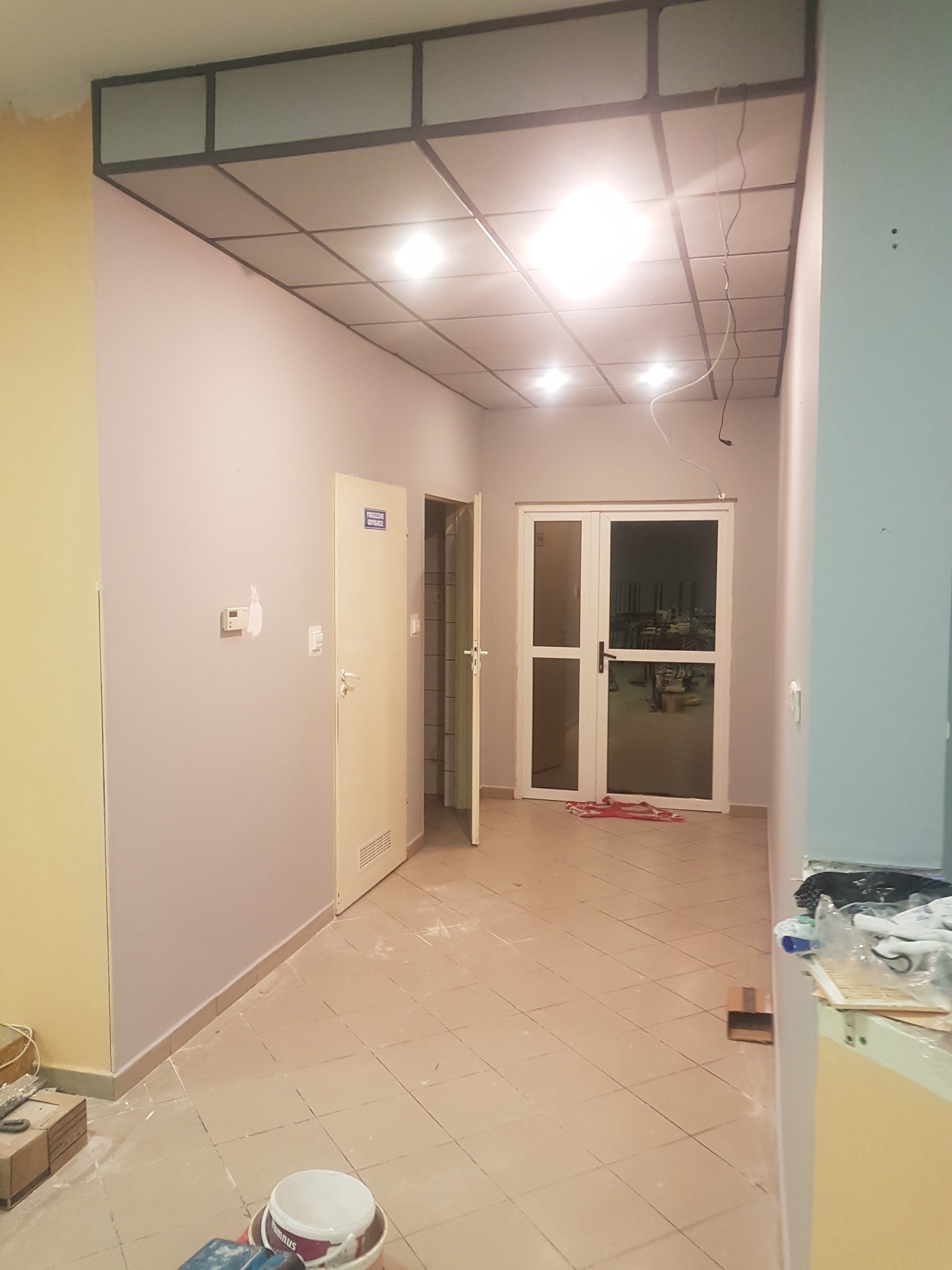 Korytarz w trakcie prac wykończeniowych: pomalowane ściany w odcieniach szarości i żółci, widoczne drzwi, sufit kasetonowy z oświetleniem, oraz akcesoria malarskie na podłodze.