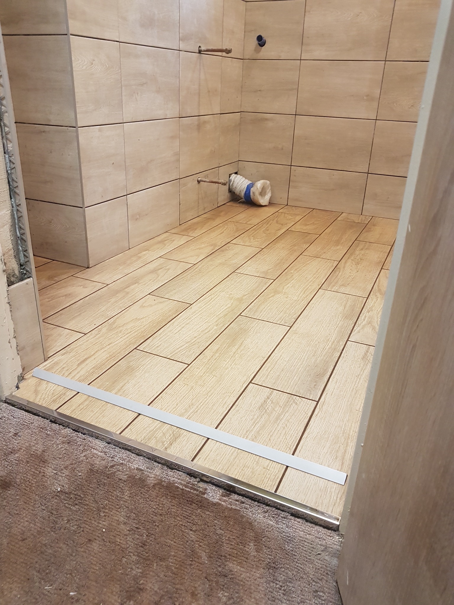 Wyłożona płytkami imitującymi drewno podłoga i ściany w trakcie remontu łazienki, widoczne niezabezpieczone rury i prowizoryczne zabezpieczenie odpływu.