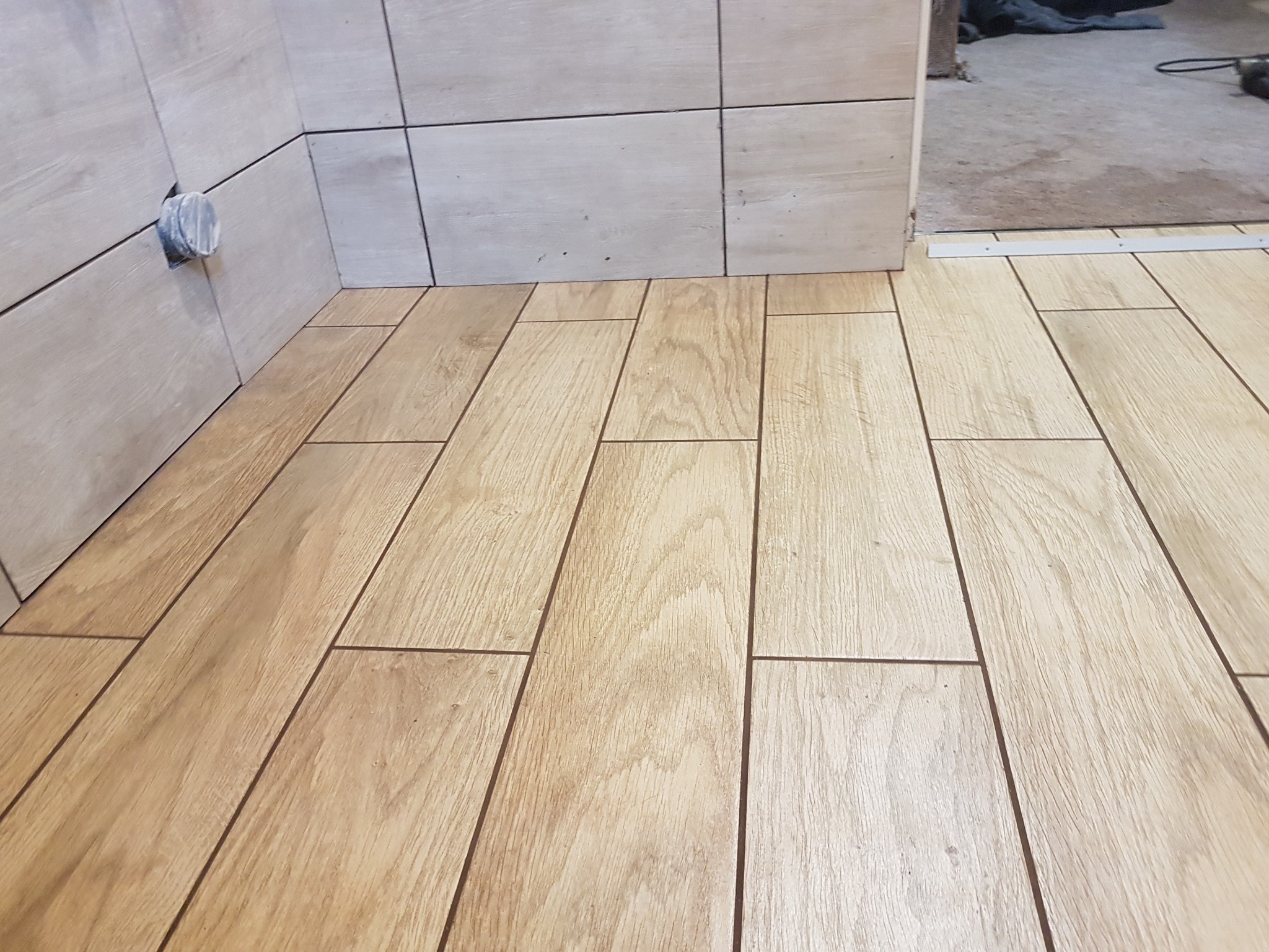 Ułożone płytki podłogowe imitujące drewno w łazience, widoczne płytki ścienne i otwór na rurę w ścianie.