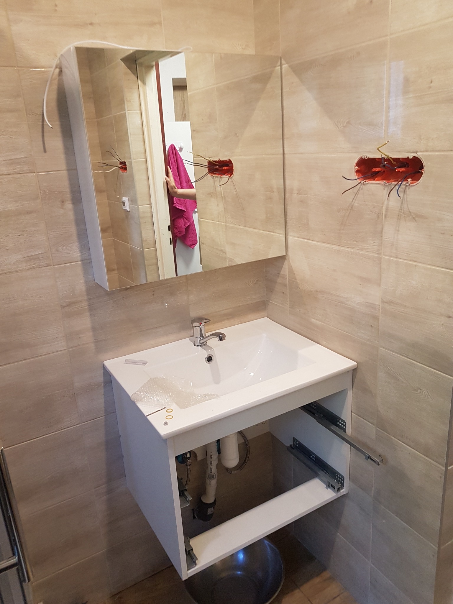 Biała umywalka z szafką i lustrem w trakcie montażu w łazience z beżowymi płytkami, widoczne wystające kable elektryczne i elementy mocujące szuflady.
