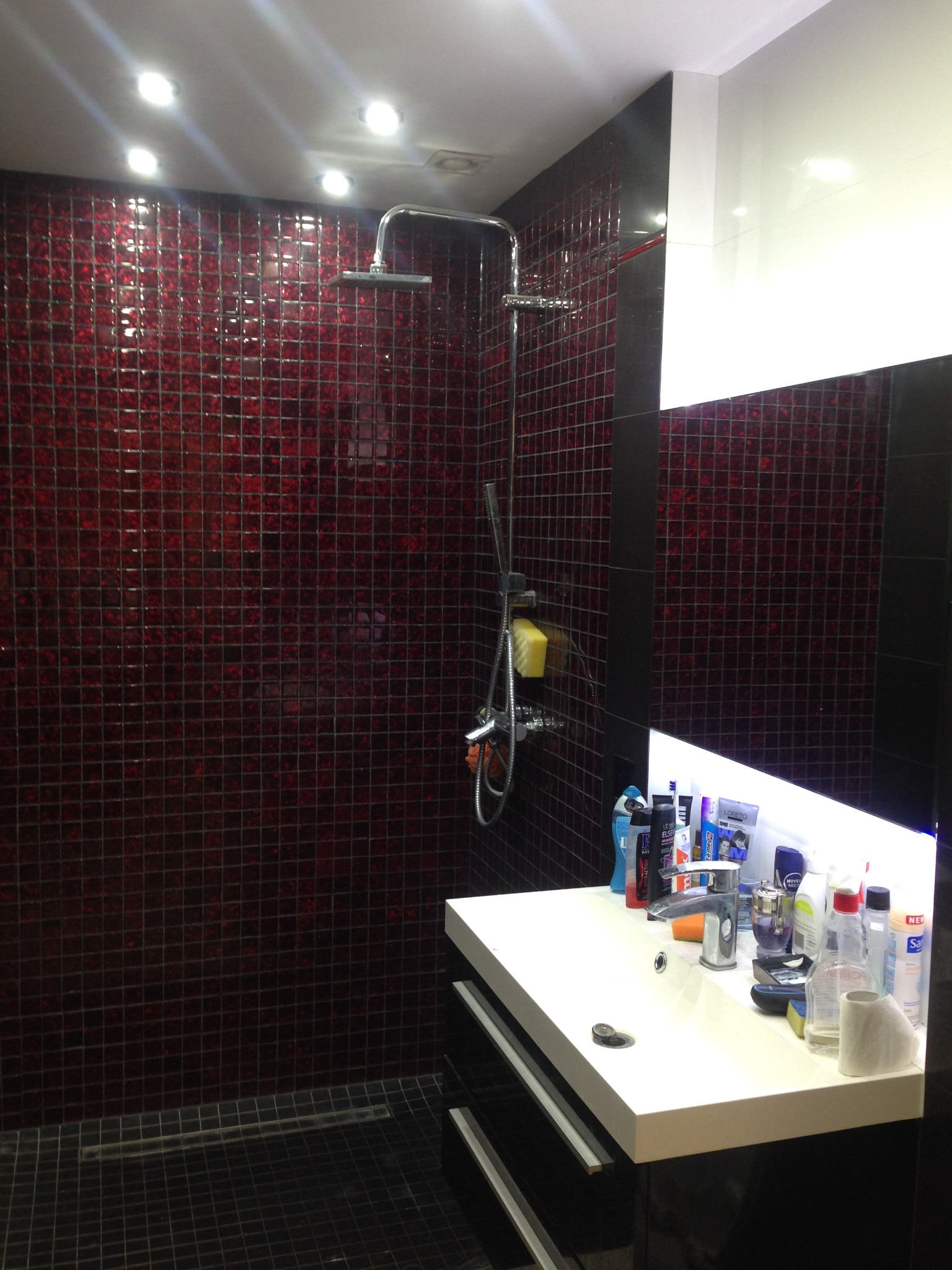 Nowoczesna łazienka z kabiną prysznicową wyłożoną bordową mozaiką, czarne płytki podłogowe, biała umywalka z czarną szafką i licznymi kosmetykami, oświetlenie punktowe.