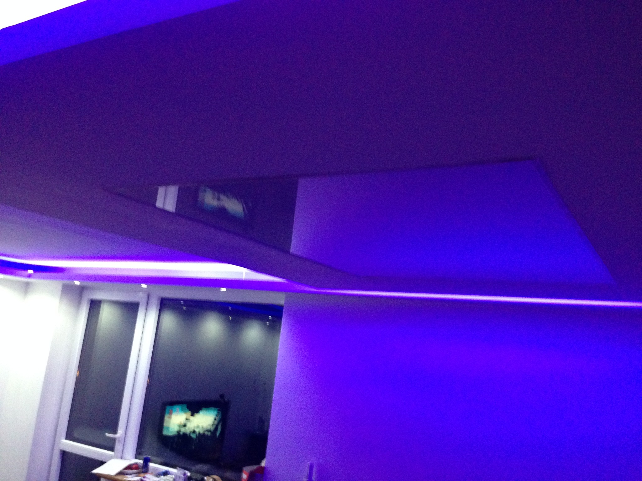 Nowoczesny sufit wielopoziomowy z podświetleniem LED w odcieniach fioletu, widok z dołu, fragment okna i ściany w tle.