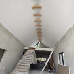 Wnętrze w trakcie prac wykończeniowych: widok na sufit z zamontowanymi drewnianymi elementami konstrukcyjnymi pod przyszłe oświetlenie, częściowo zakryte folią schody prowadzące na antresolę,...