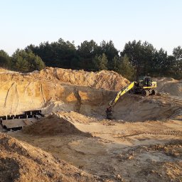 Kopczyk Piotr Strzelczyk - Żółta koparka na placu budowy wykopu pod fundamenty, widoczne szalunki i paleta z materiałami budowlanymi, w tle las iglasty i wczesne poranne światło.