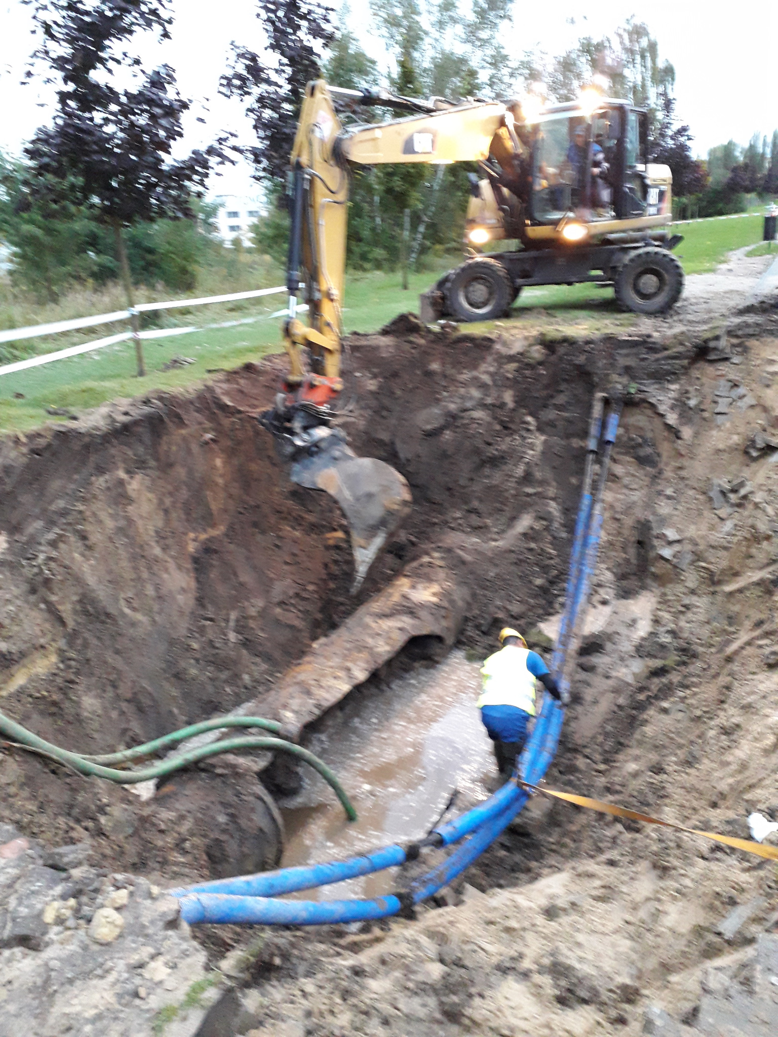 Żółta koparka CAT na skraju głębokiego wykopu z pracownikiem w kamizelce, rury i woda w dole, teren zielony w tle.