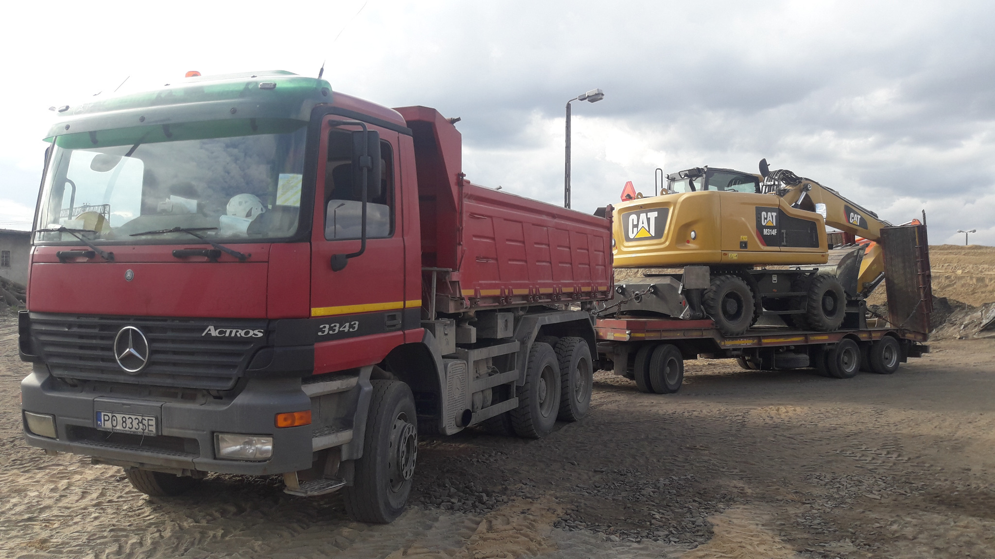 Czerwona ciężarówka Mercedes Actros 3343 z otwartą skrzynią ładunkową i żółta koparka kołowa CAT M314F na niskopodwoziowej przyczepie, stojące na placu budowy z widoczną hałdą ziemi w tle.