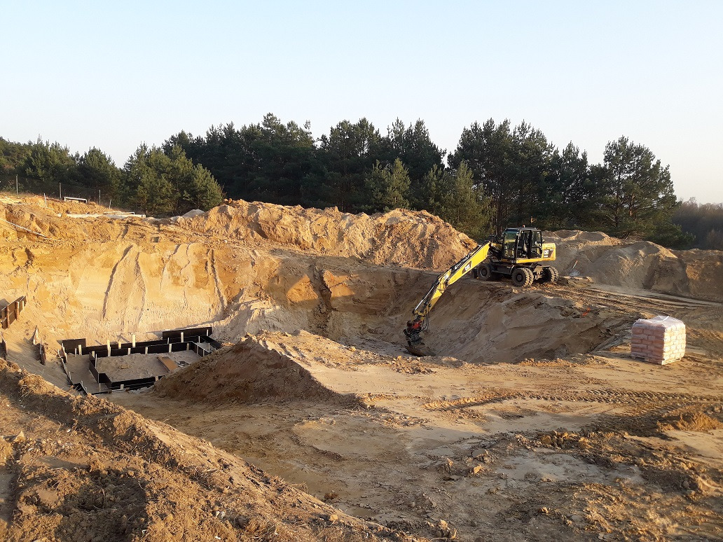 Żółta koparka na placu budowy wykopu pod fundamenty, widoczne szalunki i paleta z materiałami budowlanymi, w tle las iglasty i wczesne poranne światło.