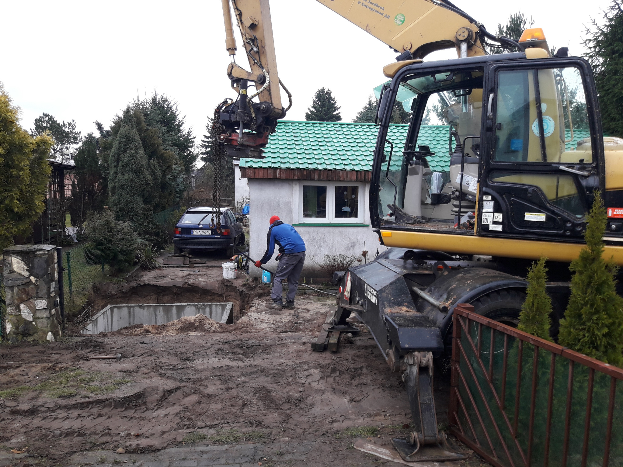 Żółty koparko-ładowarka na błotnistym terenie obok wykopu pod fundamenty, pracownik z łopatą w pobliżu, w tle budynek z zielonym dachem i zaparkowany samochód