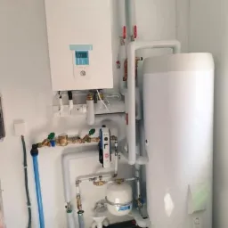 Instalacja pompy ciepła Panasonic z zasobnikiem w kotłowni w Prądkach, widoczne białe rury i osprzęt hydrauliczny.