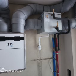 ToNat Tomasz Kozioł - Centrala rekuperacyjna IZZI zamontowana na ścianie w surowym stanie, widoczne szare rury wentylacyjne, czarne rury instalacji wodnej i niebiesko-czerwone rury CO.