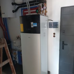 ToNat Tomasz Kozioł - Nowoczesna instalacja grzewcza z pompą ciepła Daikin, widoczne rury w kolorach niebieskim i czerwonym, zbiornik Ferolli, skrzynka elektryczna i drabina oparta o ścianę.