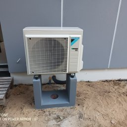 ToNat Tomasz Kozioł - Biała jednostka zewnętrzna klimatyzacji Daikin zamontowana na szarym betonowym postumencie, widoczny czarny przewód odprowadzający skropliny i paleta drewniana z boku, tło stanowi szara ściana...