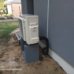 ToNat Tomasz Kozioł - Zewnętrzna jednostka klimatyzacji Daikin zamontowana na betonowej podstawie przy szarej ścianie budynku, widoczny czarny przewód chłodniczy.