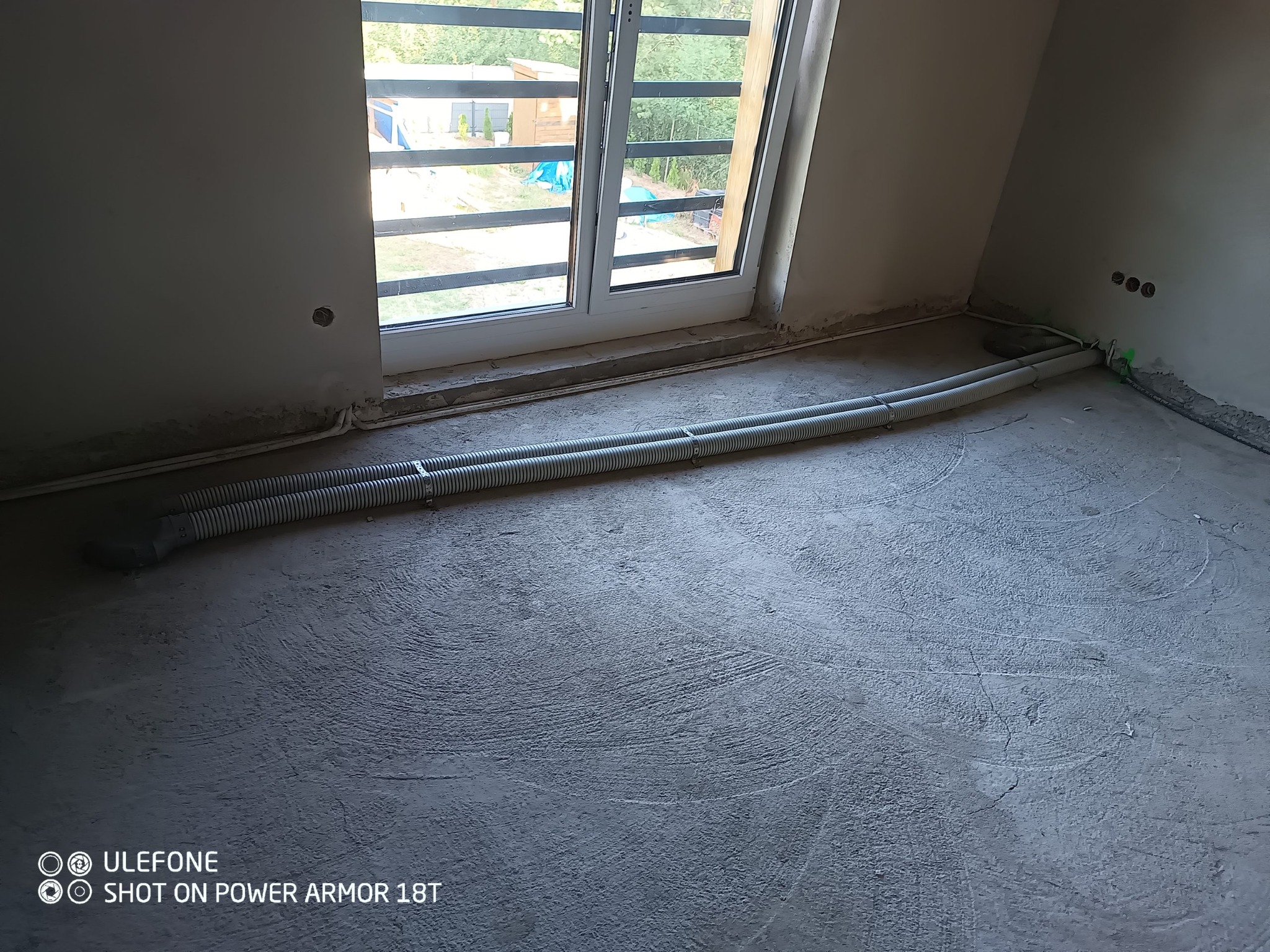 Dwa karbowane przewody wentylacyjne leżące na surowym betonowym podłożu w pomieszczeniu z oknem, widoczne przygotowane otwory w ścianie i listwy przypodłogowe.