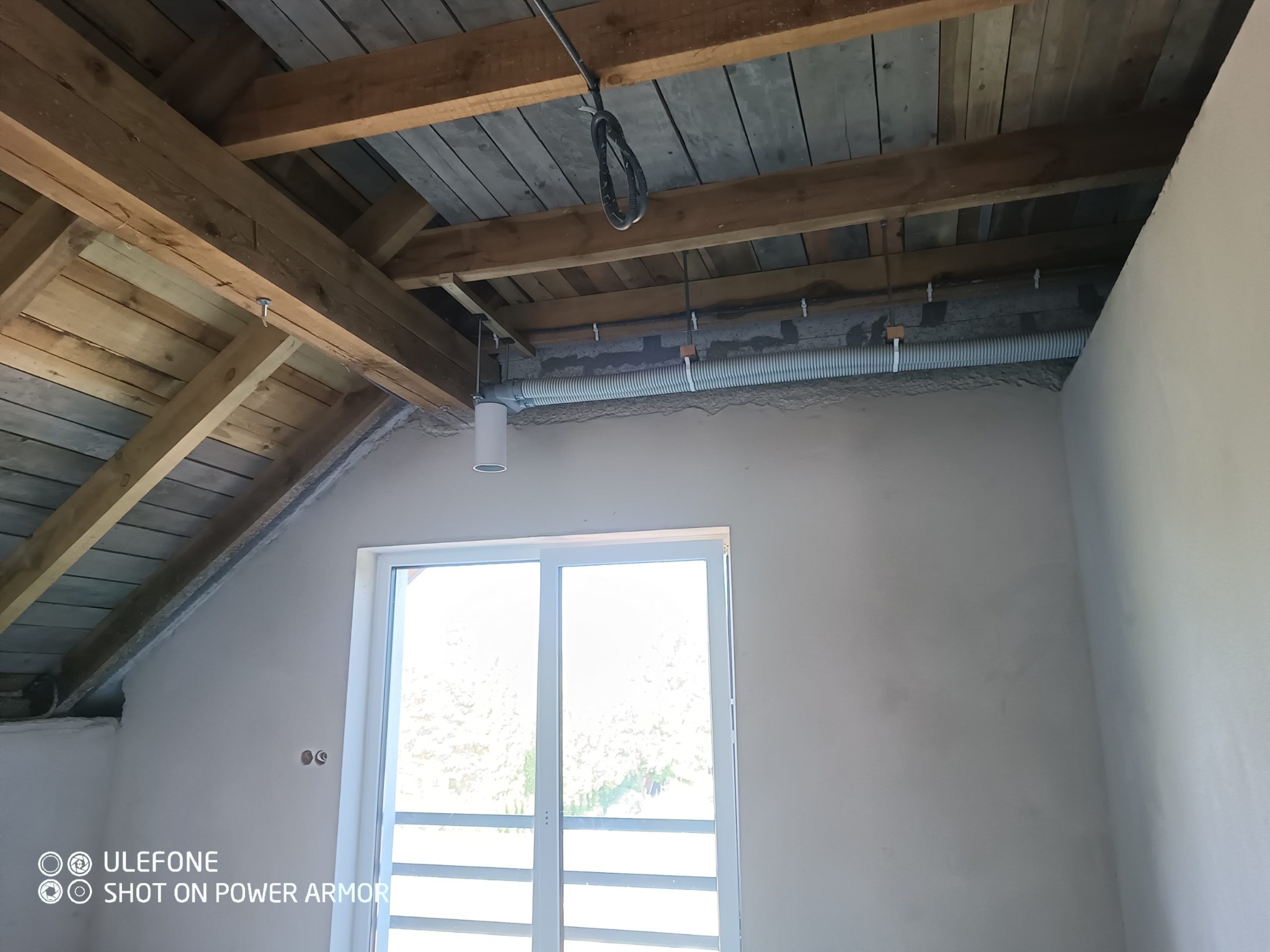 Poddasze z drewnianą konstrukcją dachu, oknem i zamontowaną rurą wentylacyjną, widoczne przewody elektryczne i niedokończone ściany.