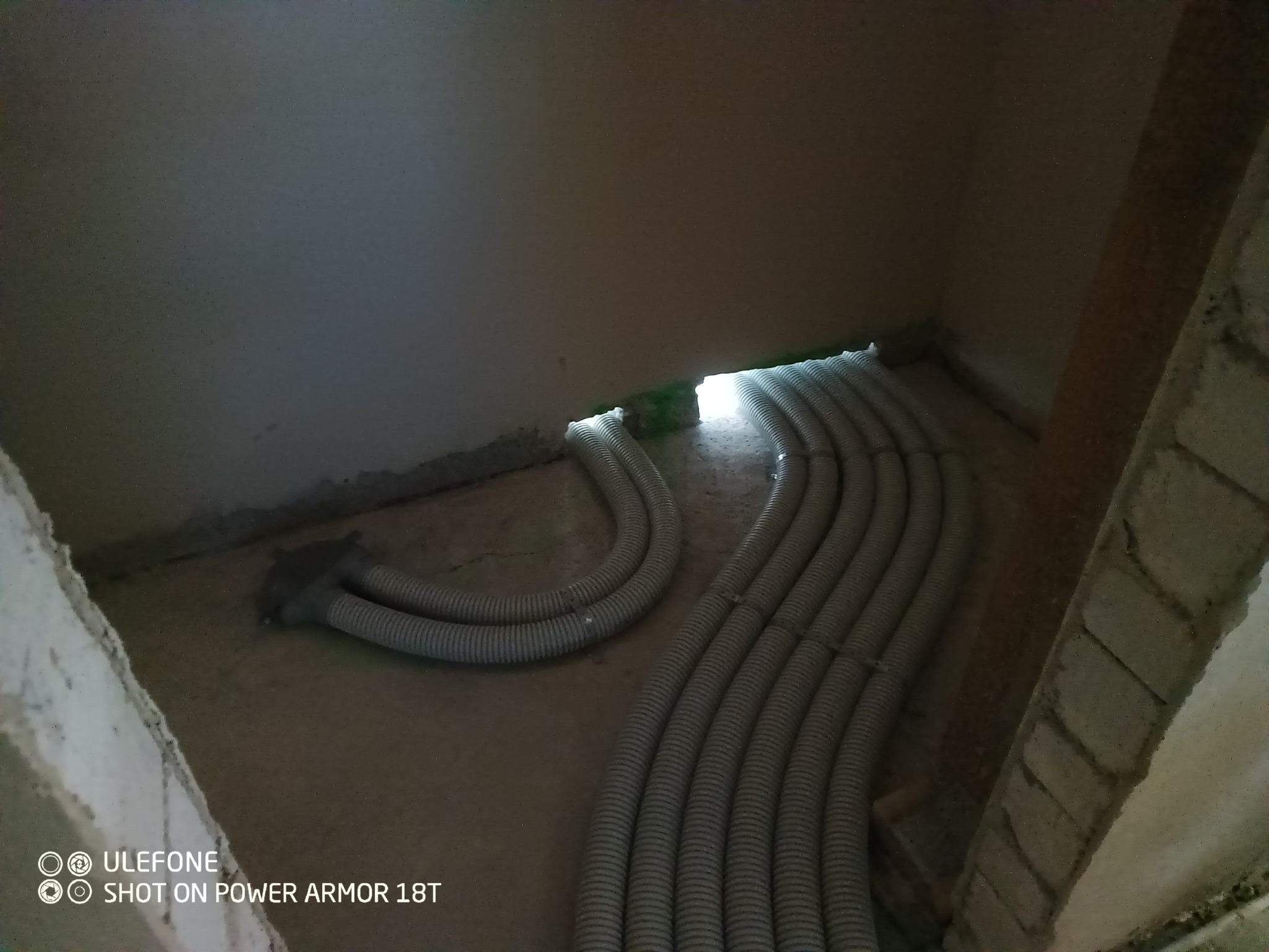 Szereg elastycznych przewodów wentylacyjnych ułożonych równolegle na surowej podłodze w trakcie montażu, widoczne fragmenty ścian w stanie deweloperskim, oświetlenie z góry.